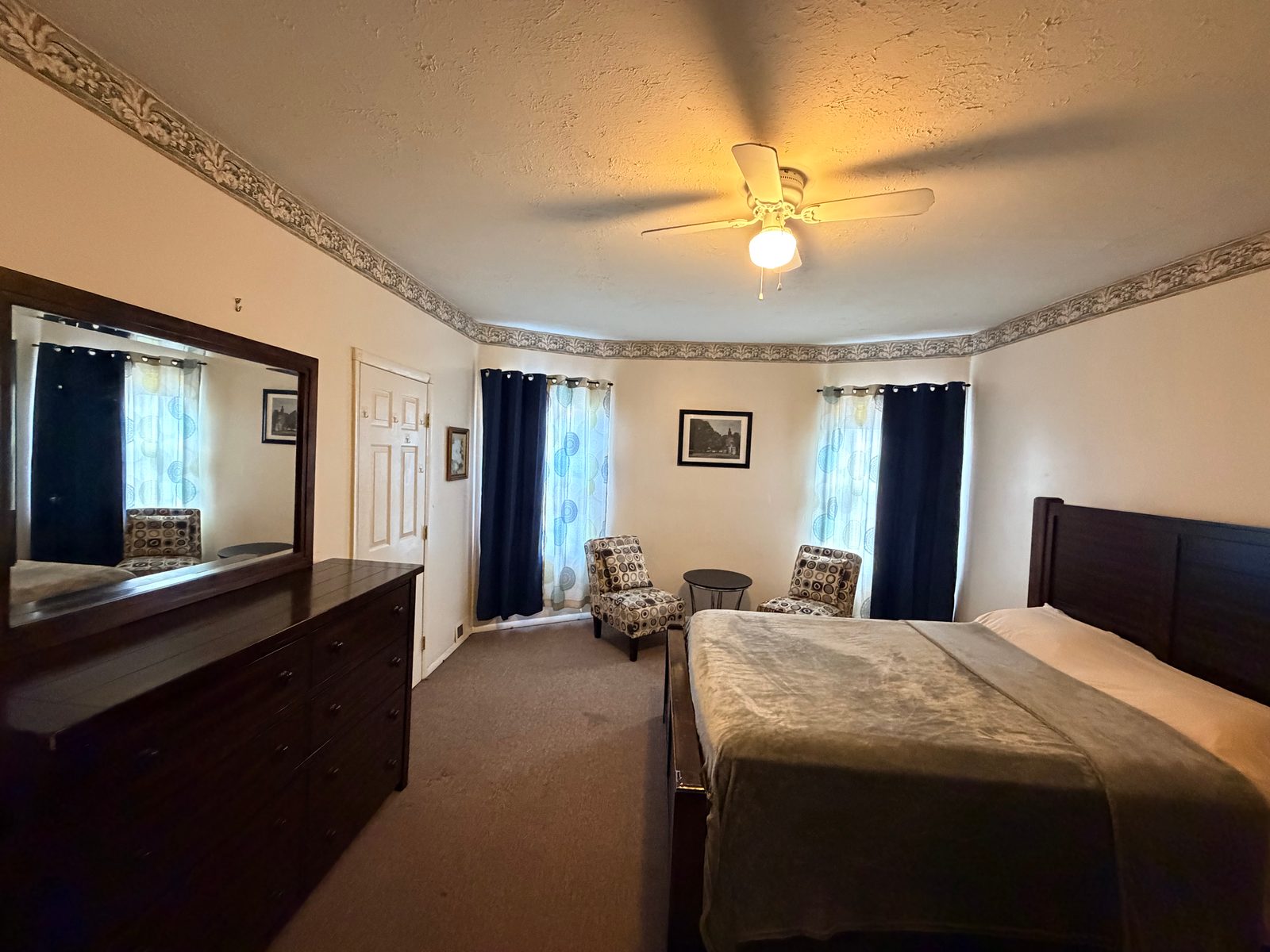 Furnished private bedroom with bed, dresser, mirror, seating, and windows